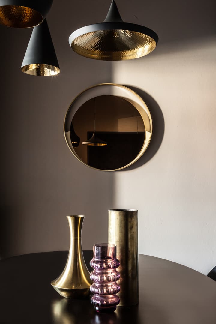A modern interior with a round mirror, three pendant lights, and a table holding gold and purple vases. Light casts shadows on the wall.