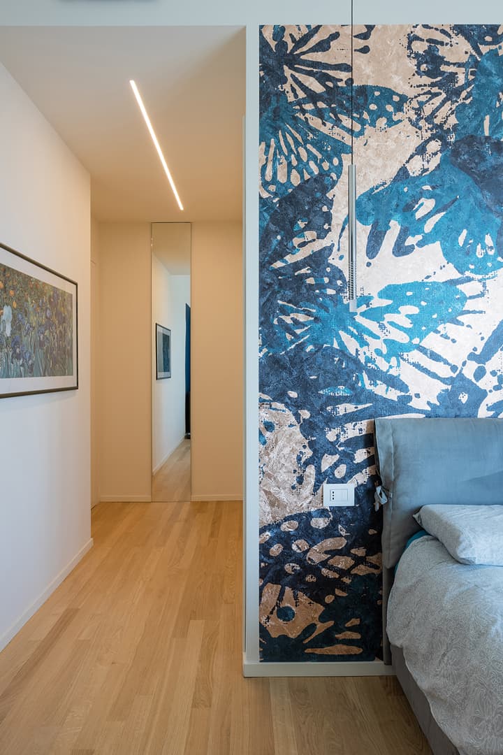 Bedroom with a bed, blue floral accent wall, wooden floor, and a hallway with artwork and a large mirror.