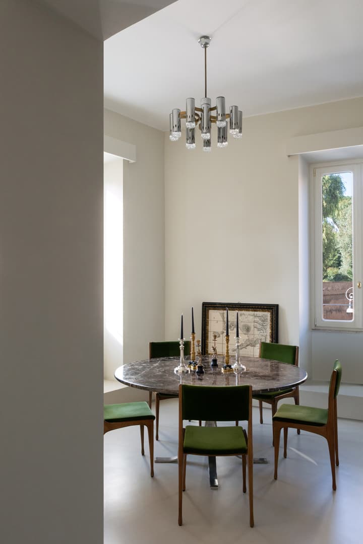 A modern dining room with a round table, green chairs, a silver chandelier, and a large window.