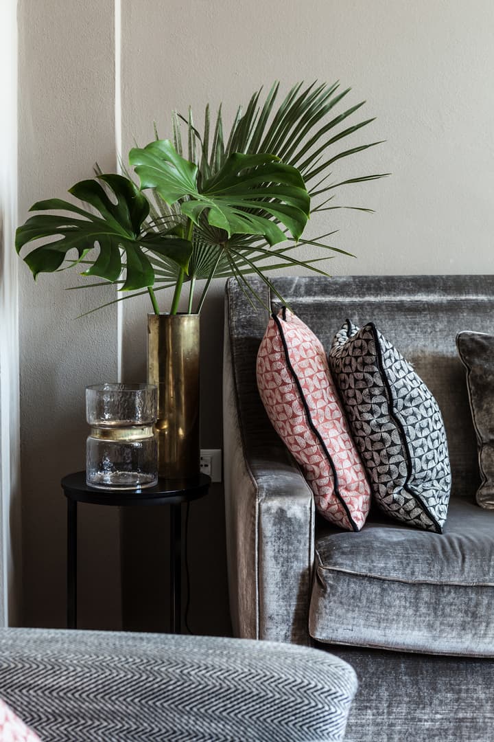 Elegant living room with a gray velvet sofa, patterned cushions, a brass vase with green leaves, and a small black side table with glass decor.