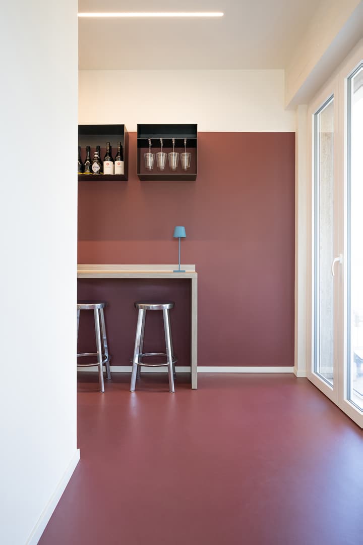 Minimalist room with bordeaux walls and floor, a small bar with two stools, a shelf with bottles and glasses, and a blue lamp.