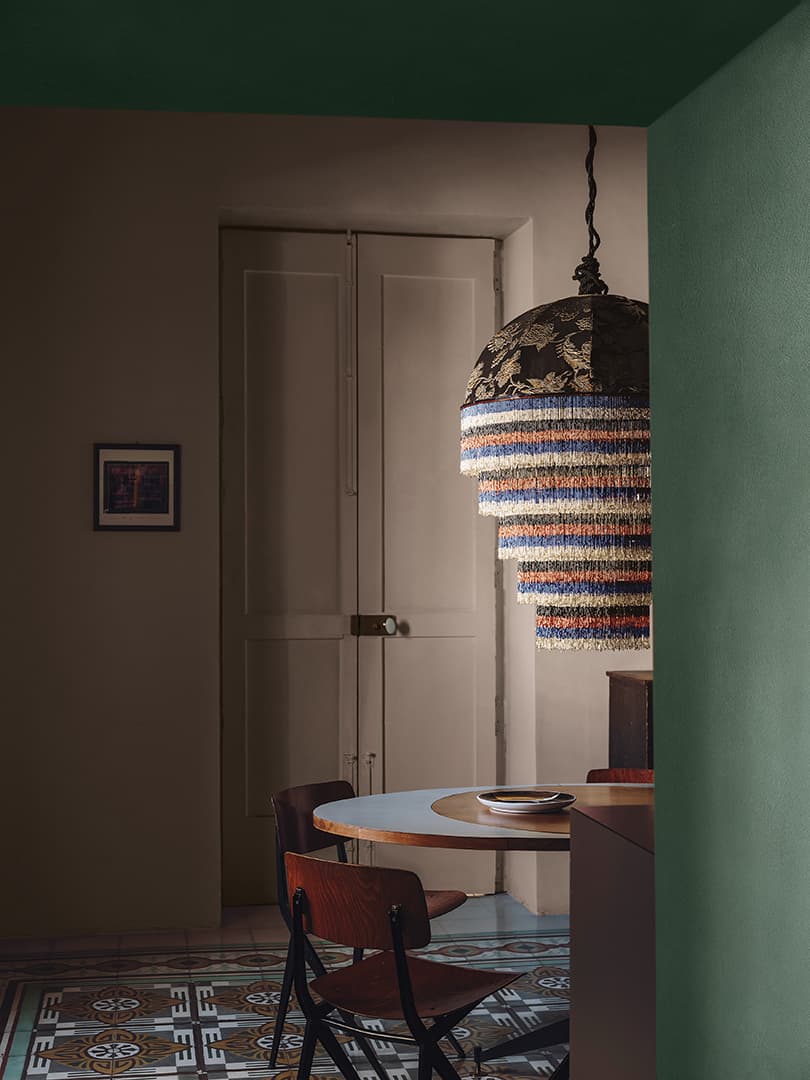 Cozy room with a round table, wooden chairs, and a colorful fringe chandelier. Soft lighting highlights the patterned floor and closed doors.