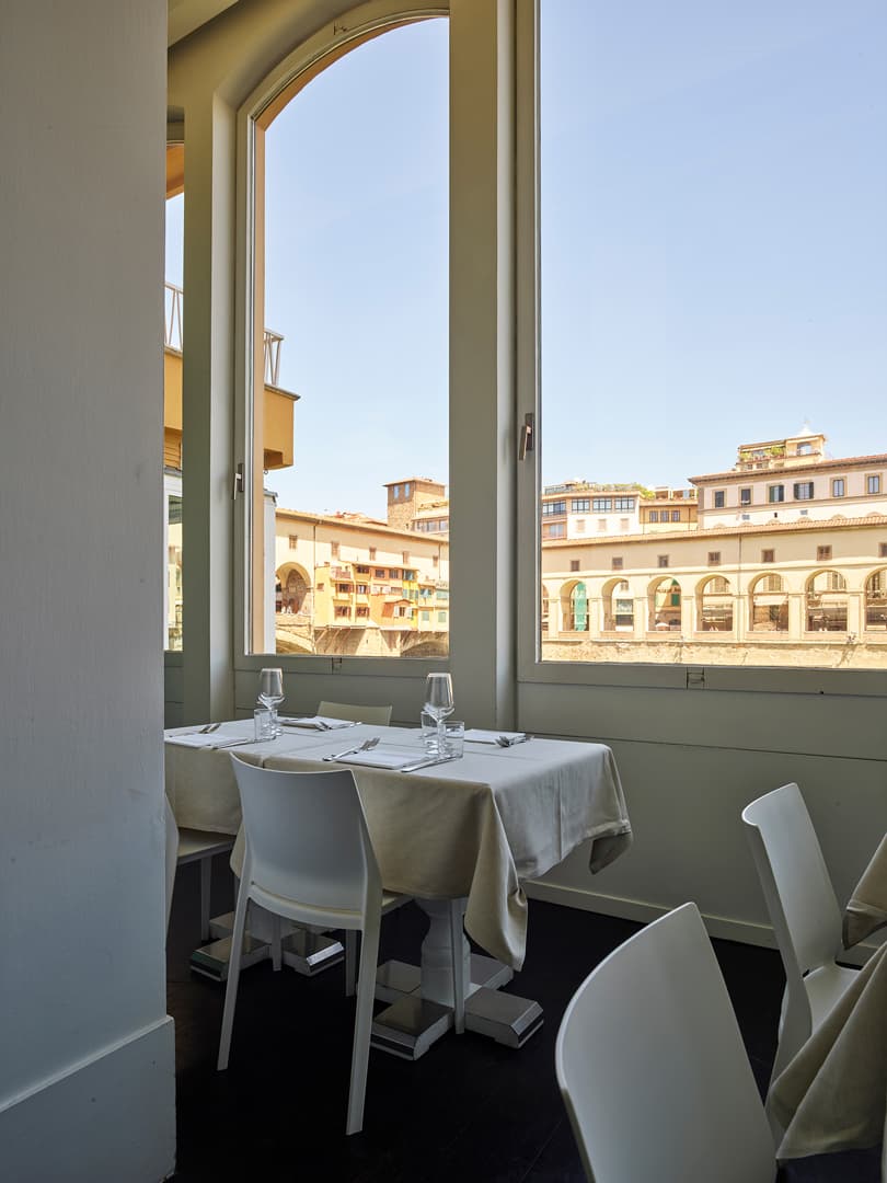 A sunlit restaurant interior with white tables and chairs, large windows offering a view of historic buildings under a clear blue sky.