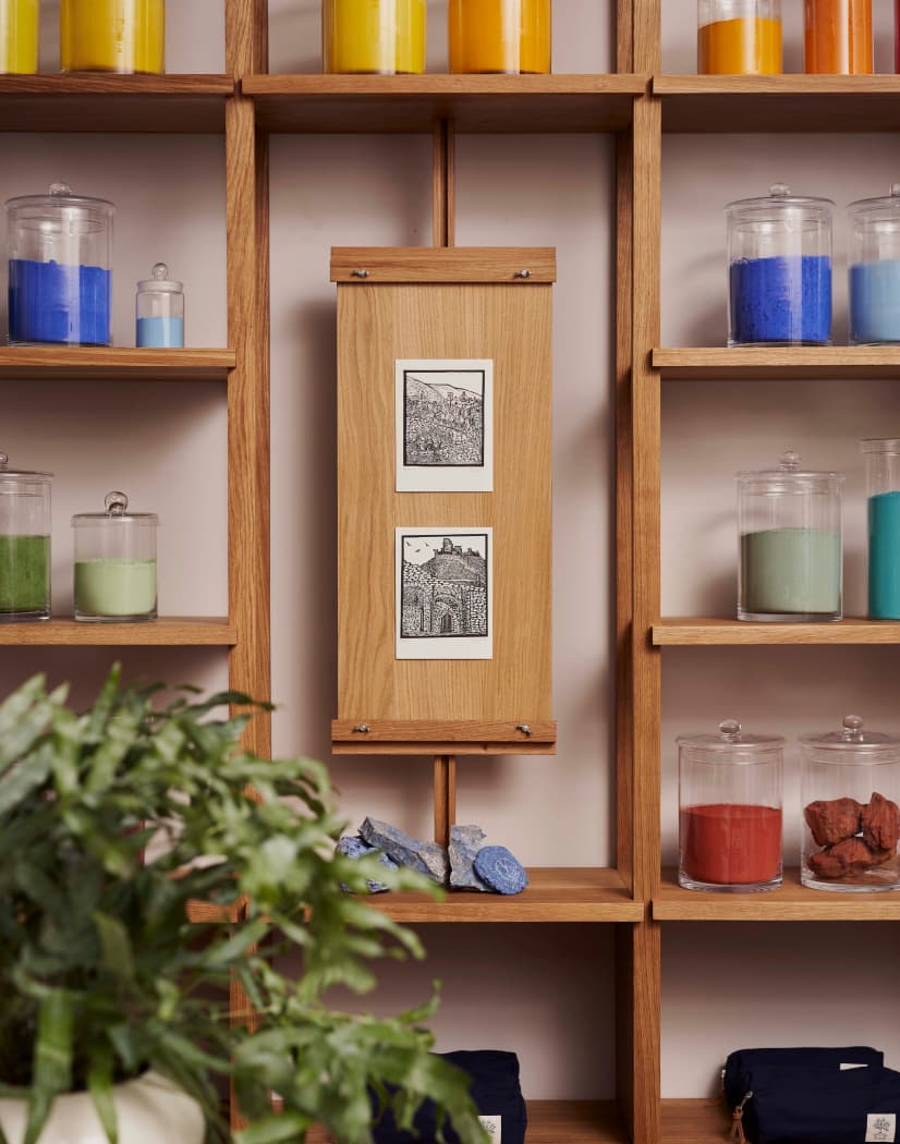 Wooden shelves with jars of colorful pigments and a framed artwork in the center. A plant is partially visible in the foreground.